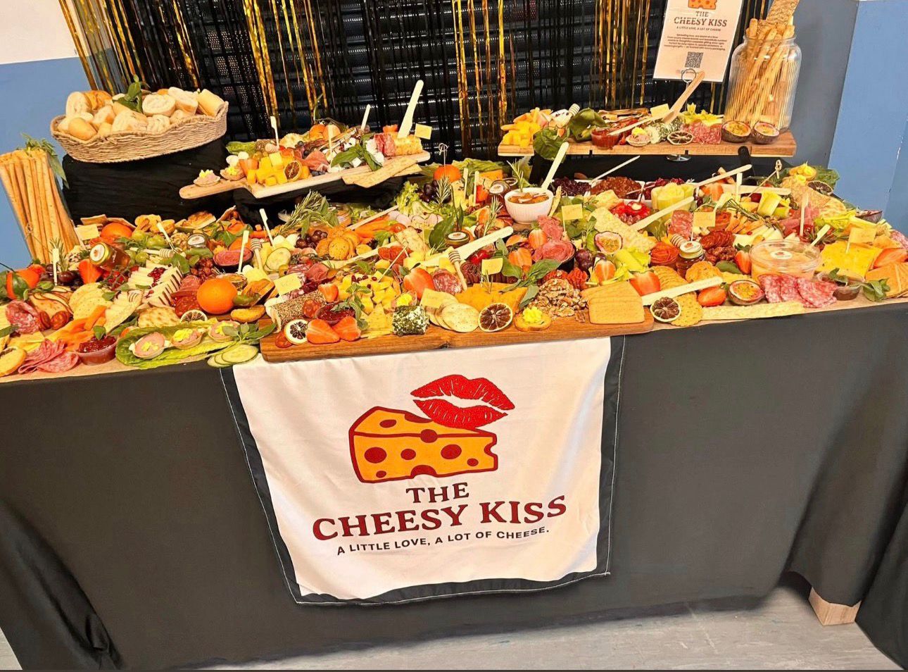 Full-length Cheesy Kiss grazing table with gold backdrop and branded napkin